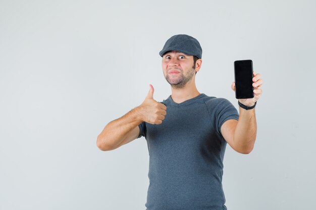 Young male in t-shirt cap showing thumb up while holding mobile phone  