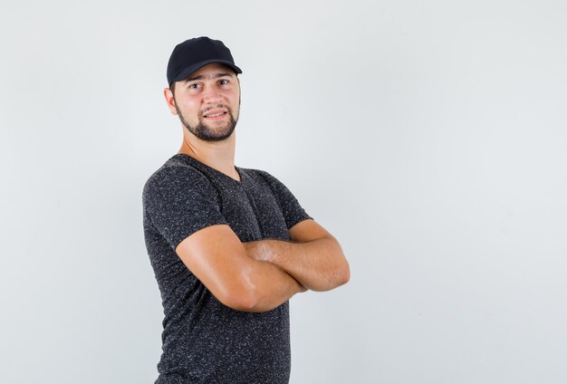 Young male in t-shirt and cap standing with crossed arms and looking cheerful