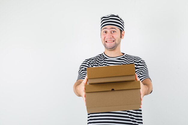 Young male in t-shirt, hat delivering cardboard boxes and looking cheery , front view.