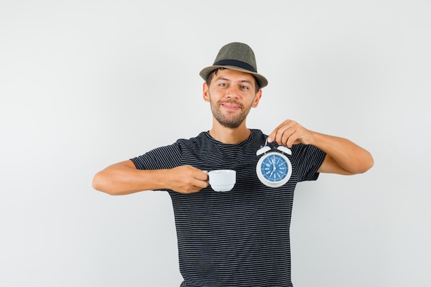 Foto gratuita giovane maschio in cappello della maglietta che tiene sveglia e tazza di bevanda e che sembra contento