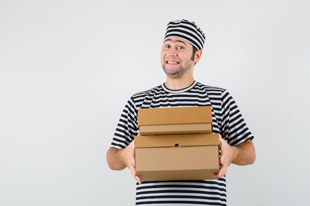 Young male in t-shirt, hat holding cardboard boxes and looking jolly , front view.