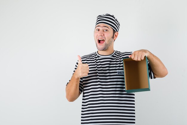Молодой мужчина в футболке, шляпе держит пустую подарочную коробку, показывает палец вверх и выглядит счастливым, вид спереди.