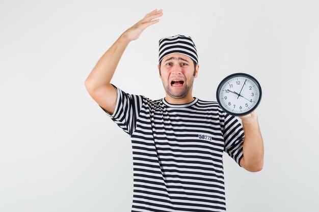 Young male in t-shirt, hat holding wall clock and looking wistful , front view.