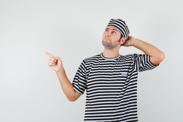 Young male in t-shirt, hat pointing aside and looking hesitant , front view.