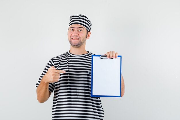 Young male in t-shirt, hat pointing at clipboard and looking cheerful , front view.