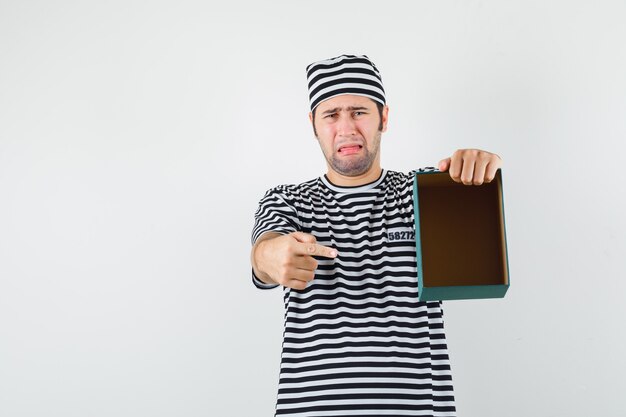 Young male in t-shirt, hat pointing at empty gift box and looking downcast , front view.