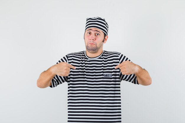 Young male in t-shirt, hat pointing at himself and looking perplexed , front view.