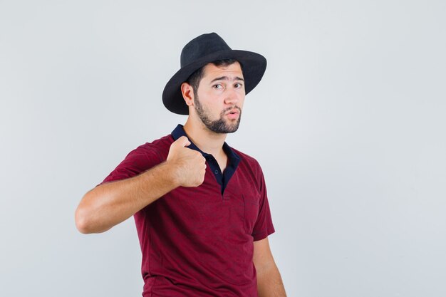 Молодой мужчина в футболке, шляпа указывает на себя и выглядит неудобно, вид спереди.