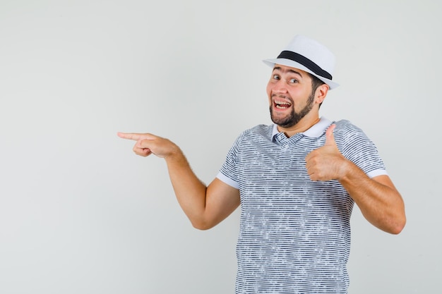 Young male in t-shirt, hat pointing to the side with thumb up and looking optimistic , front view.