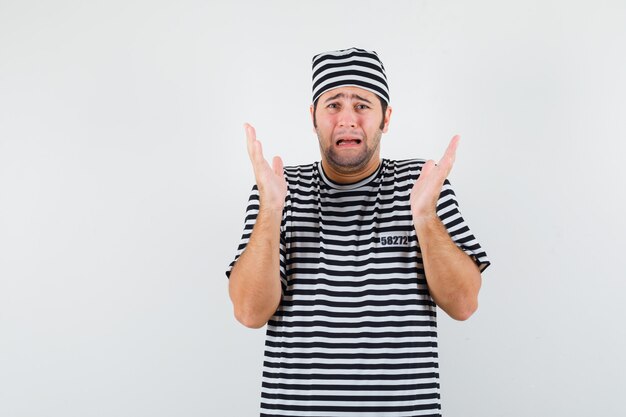 Young male in t-shirt, hat raising hands while crying and looking mournful , front view.