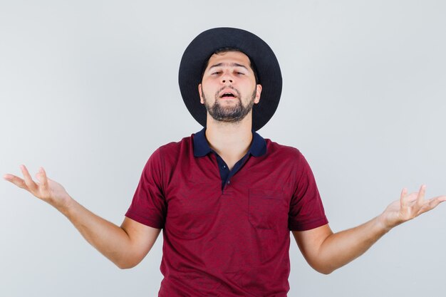 Молодой мужчина в футболке, шляпе показывает беспомощный жест и выглядит недовольным, вид спереди.