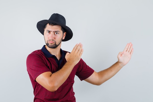 Молодой мужчина в футболке, шляпе показывает жест отбивной каратэ и выглядит сердитым, вид спереди.