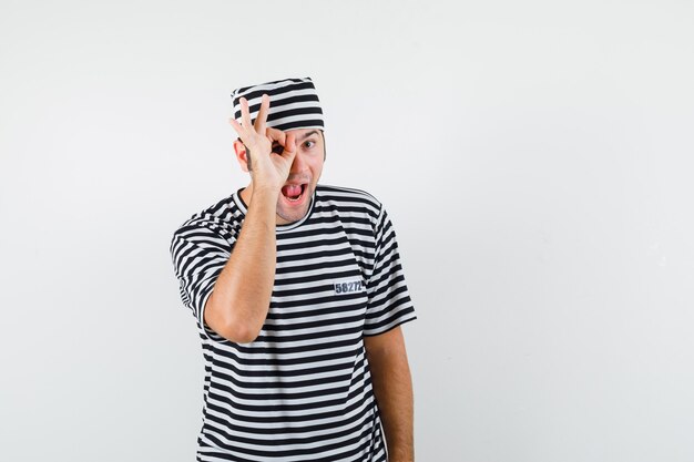 Young male in t-shirt, hat showing ok sign on eye and looking glad , front view.