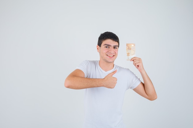 Молодой мужчина в футболке держит евробанкноту, показывает палец вверх и выглядит довольным, вид спереди.