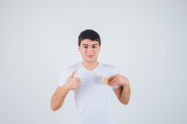 Молодой мужчина в футболке держит евробанкноту, показывает палец вверх и выглядит довольным, вид спереди.