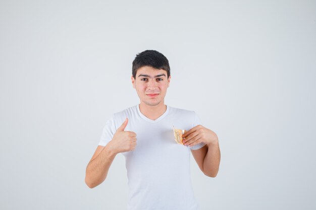 Young male in t-shirt holding eurobanknote, showing thumb up and looking pleased , front view.