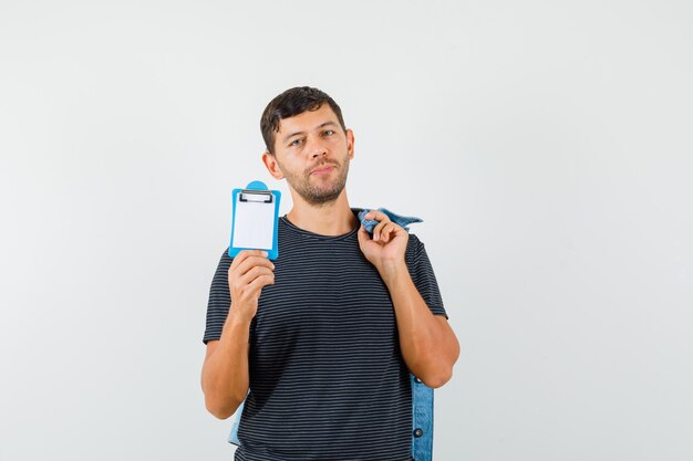 Young male in t-shirt holding jacket and mini clipboard and looking sensible  