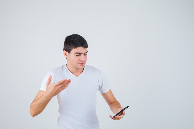 Young male in t-shirt holding phone while raising hand and looking focused , front view.