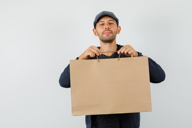 Young male in t-shirt, jacket, cap holding paper bag and smiling , front view.