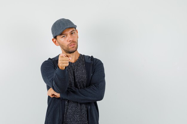 Young male in t-shirt, jacket, cap pointing at camera and looking confident , front view.