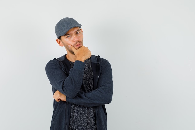 Young male in t-shirt, jacket, cap standing in thinking pose and looking intelligent , front view.