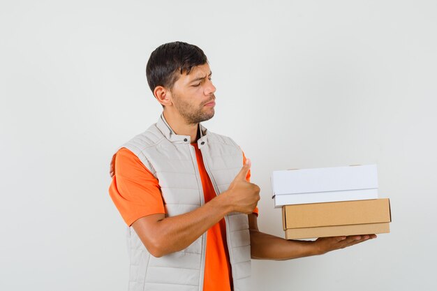 Молодой мужчина в футболке, пиджаке держит картонные коробки, показывает палец вверх, вид спереди.