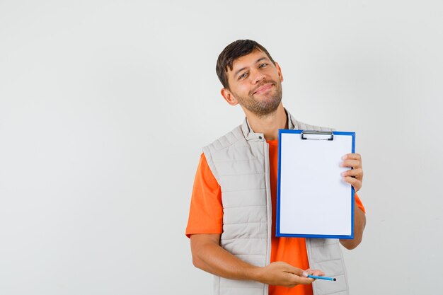 Young male in t-shirt, jacket holding clipboard and pencil and looking merry , front view.