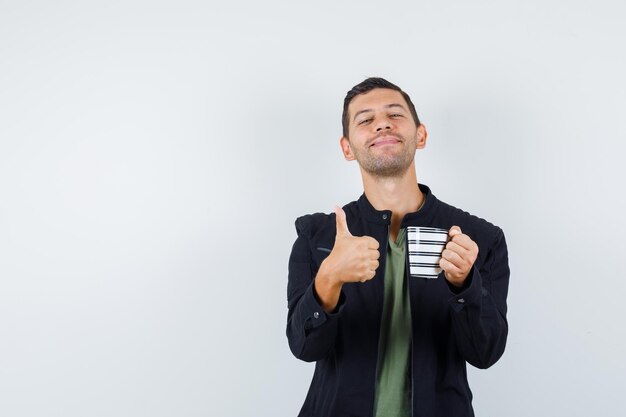 Young male in t-shirt, jacket holding cup of drink with thumb up and looking cheery , front view.