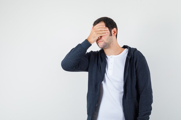 Young male in t-shirt, jacket holding hand on face and looking forgetful , front view.