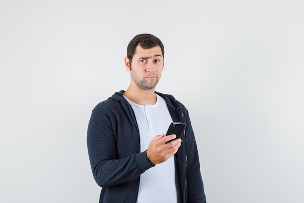 Молодой мужчина в футболке, куртке, держащей мобильный телефон и нерешительной, вид спереди.