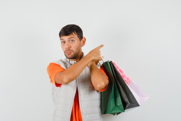 Young male in t-shirt, jacket holding shopping bags, pointing away , front view.