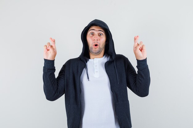 Young male in t-shirt, jacket keeping fingers crossed and looking surprised , front view.