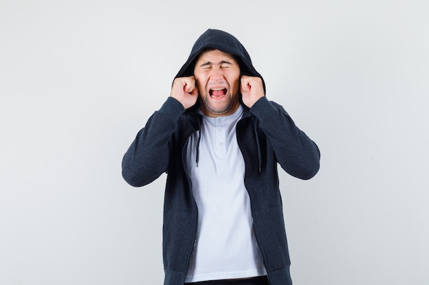 Young male in t-shirt, jacket plugging ears with fingers and looking annoyed , front view.