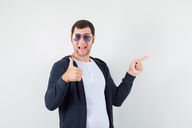 Young male in t-shirt, jacket pointing aside, showing thumb up and looking optimistic , front view.