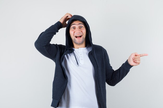 Young male in t-shirt, jacket pointing aside while scratching head and looking happy , front view.