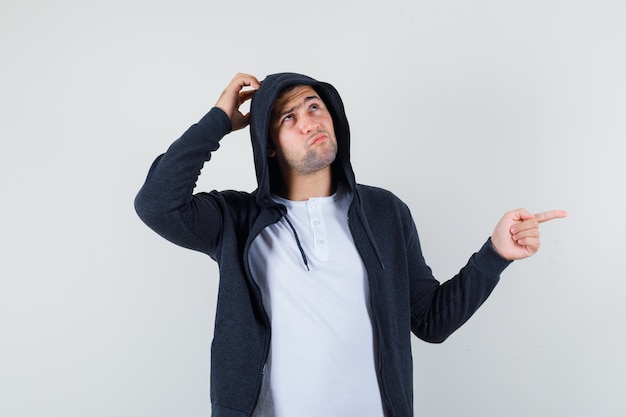 Young male in t-shirt, jacket pointing aside while scratching head and looking hesitant , front view.