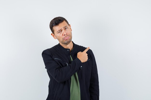 Young male in t-shirt, jacket pointing away and looking offended , front view.