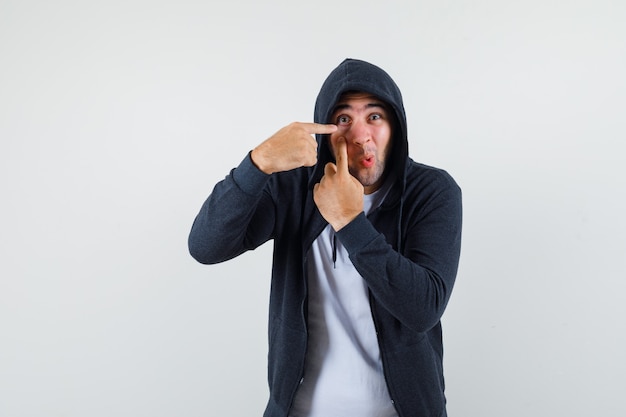 Young male in t-shirt, jacket pointing at his eye and looking funny , front view.