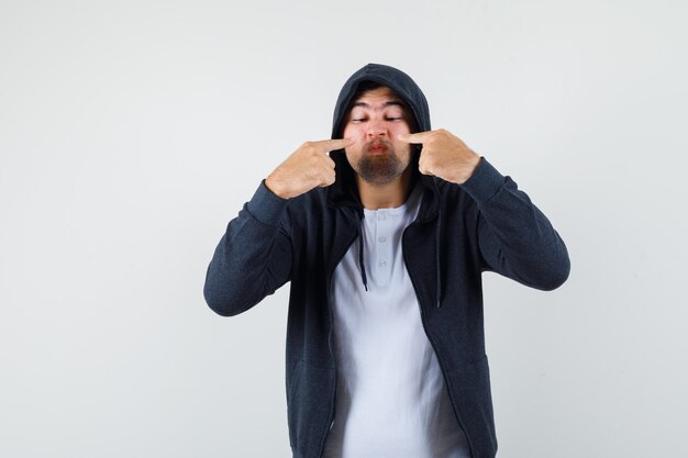 Young male in t-shirt, jacket pointing at his nose and looking focused , front view.