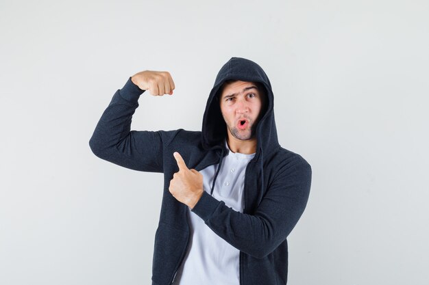 Young male in t-shirt, jacket pointing at muscles of arm and looking confident , front view.