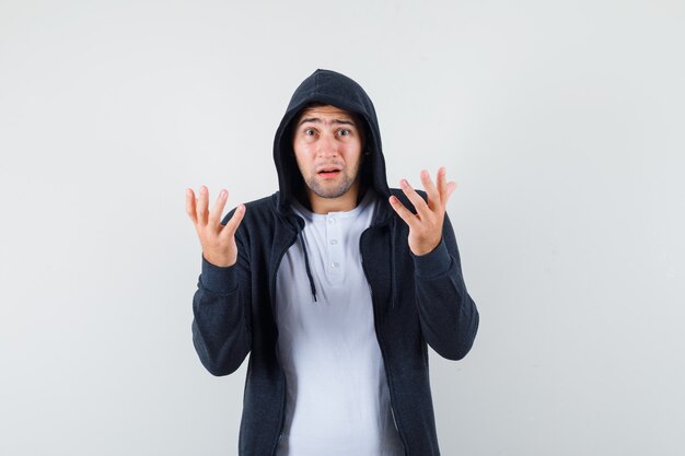 Young male in t-shirt, jacket raising hands and looking confused , front view.