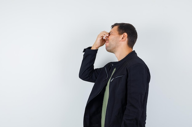 Young male in t-shirt, jacket rubbing eyes and nose and looking forgetful .