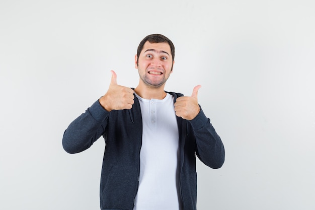 Young male in t-shirt, jacket showing double thumbs up and looking merry , front view.