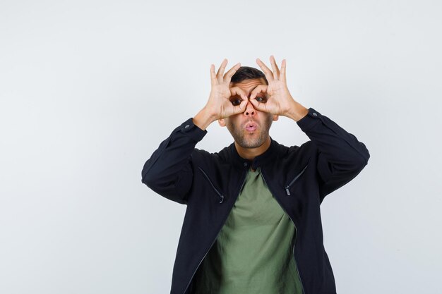 Young male in t-shirt, jacket showing glasses gesture and looking amazed , front view.