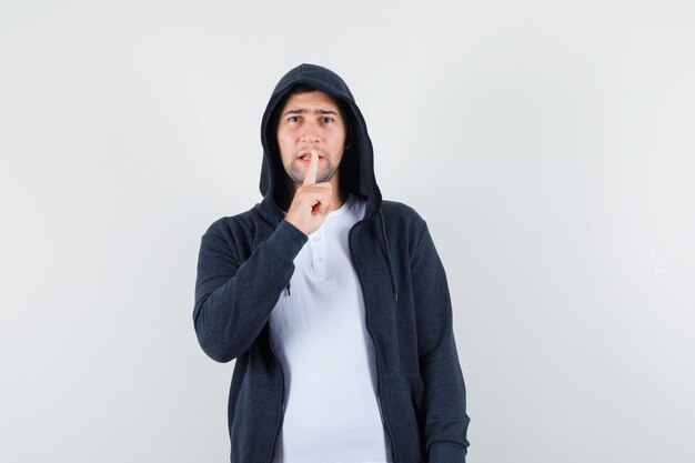 Молодой мужчина в футболке, пиджаке показывает жест молчания и внимательно смотрит, вид спереди.