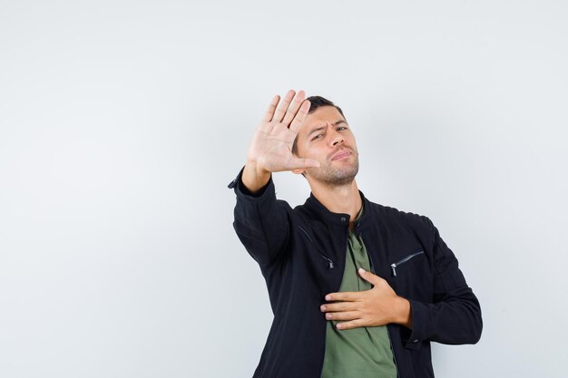 Young male in t-shirt, jacket showing stop gesture and looking bored , front view.