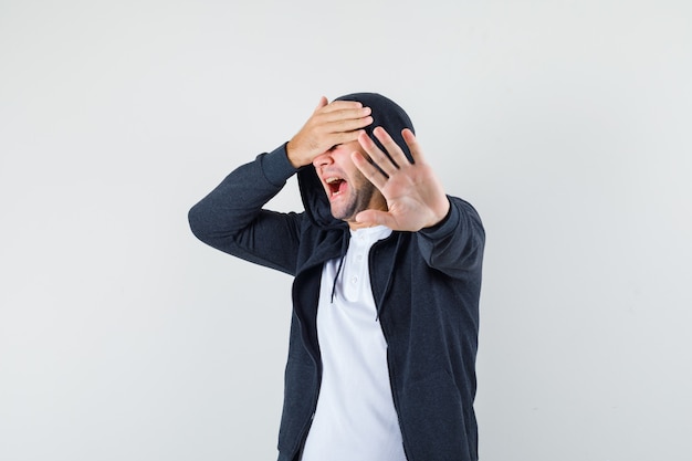 Young male in t-shirt, jacket showing stop gesture and looking frightened, front view.