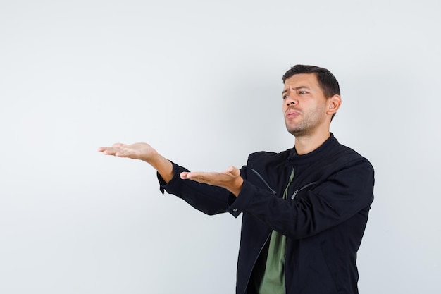 Young male in t-shirt, jacket stretching hands in disapproval gesture and looking downcast , front view.