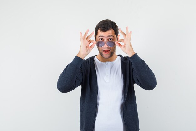 Young male in t-shirt, jacket taking off glasses and looking amazed , front view.