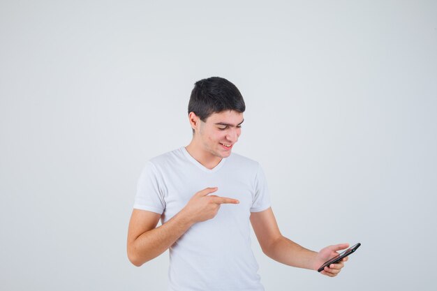 Young male in t-shirt pointing at phone and looking merry , front view.